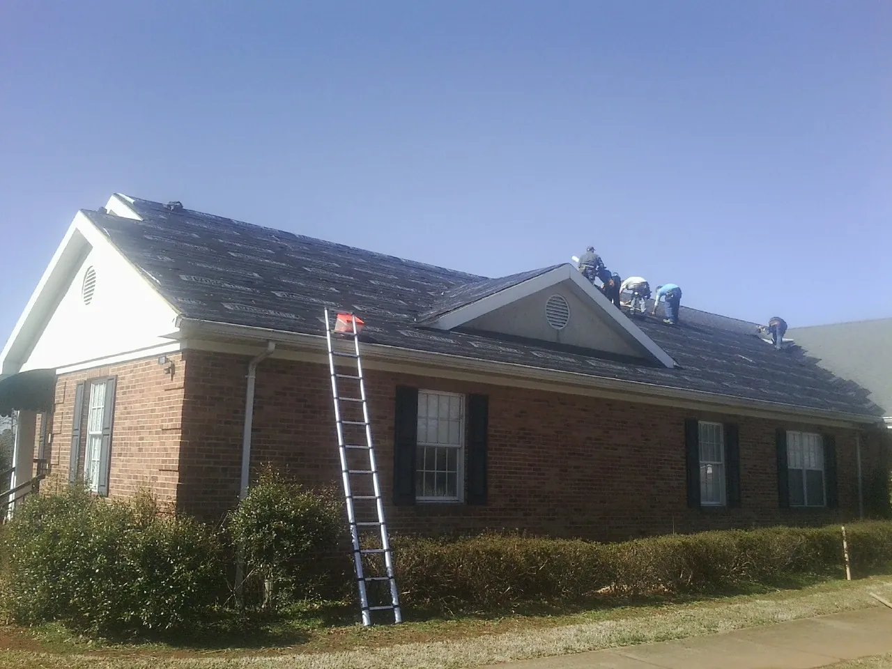 Crew tearing shingles off a brick ranch roof with a ladder set — Emergency Roof Repair work by our crew in Hematite
